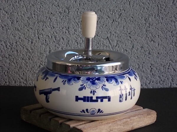 Cendrier poussoir publicitaire pour la marque "Hilti". Corps en faïence Blanche à motif Bleu de N.V Plateelbakkerij et de N.V Metawa pour le poussoir en métal. Made in Holland