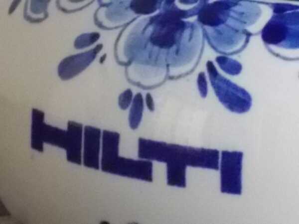 Cendrier poussoir publicitaire pour la marque "Hilti". Corps en faïence Blanche à motif Bleu de N.V Plateelbakkerij et de N.V Metawa pour le poussoir en métal. Made in Holland